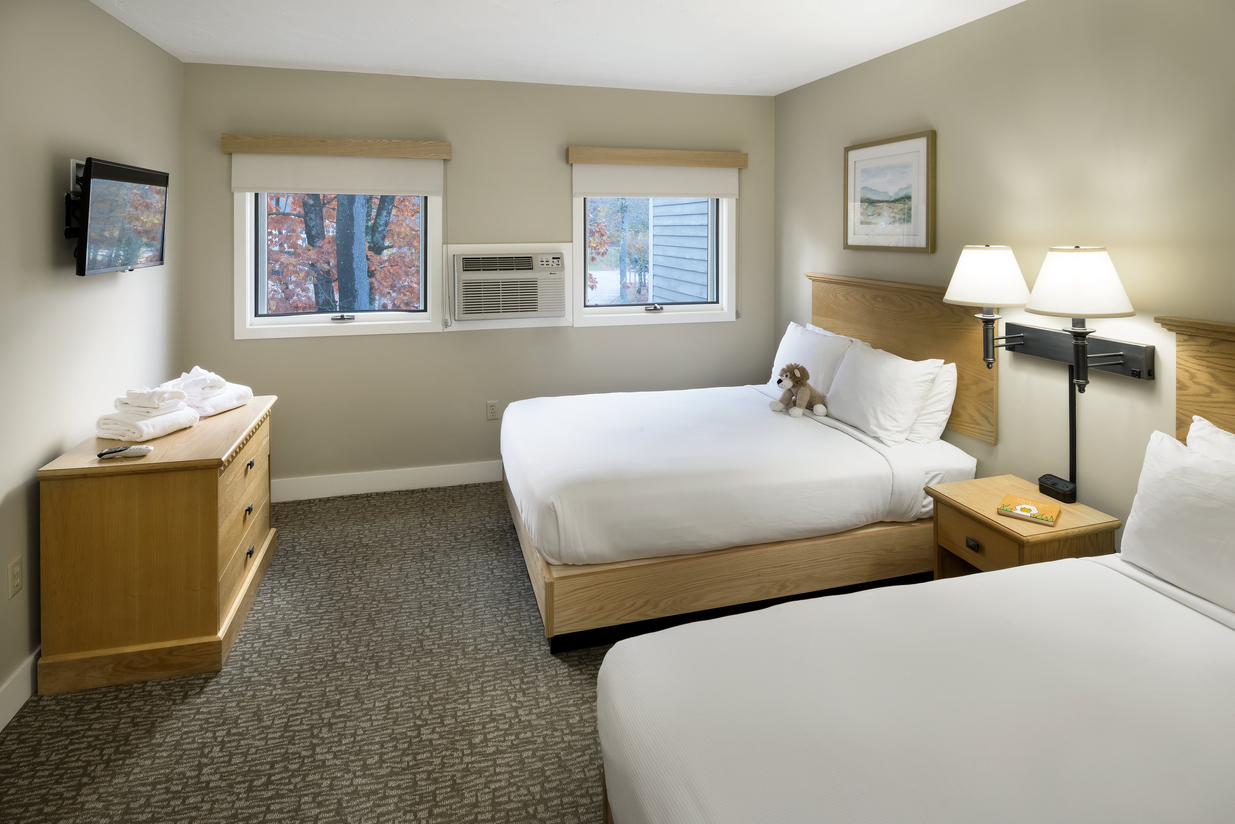 Bedroom with two queen beds, wall-mounted TV, dresser stacked with towels, and autumn trees outside