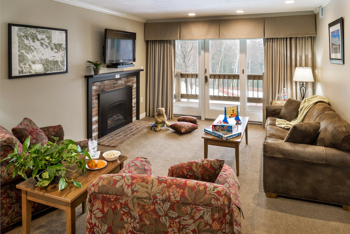 Suite living room with a brick fireplace, leather sofa, board games and a balcony door to the woods