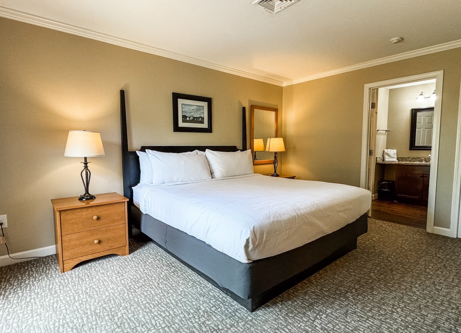 King bed with white linens between two lamps, with an open doorway to the bathroom vanity