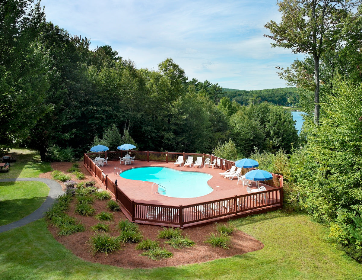 Outdoor pool on a wood deck with blue umbrellas, framed by trees above a distant lake